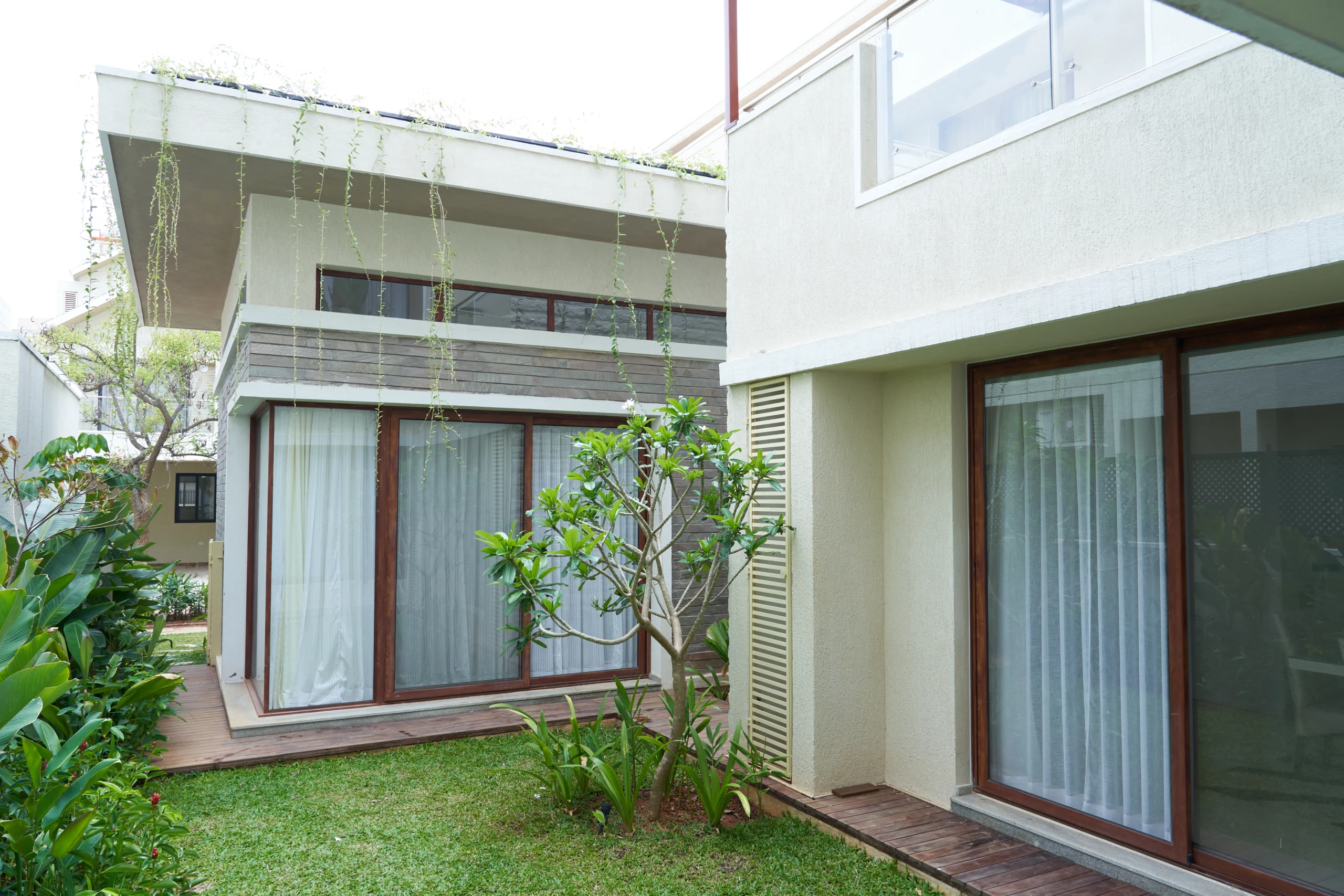 Luxury villa courtyard with lawn, wooden deck flooring, large glass windows, and tropical landscape design